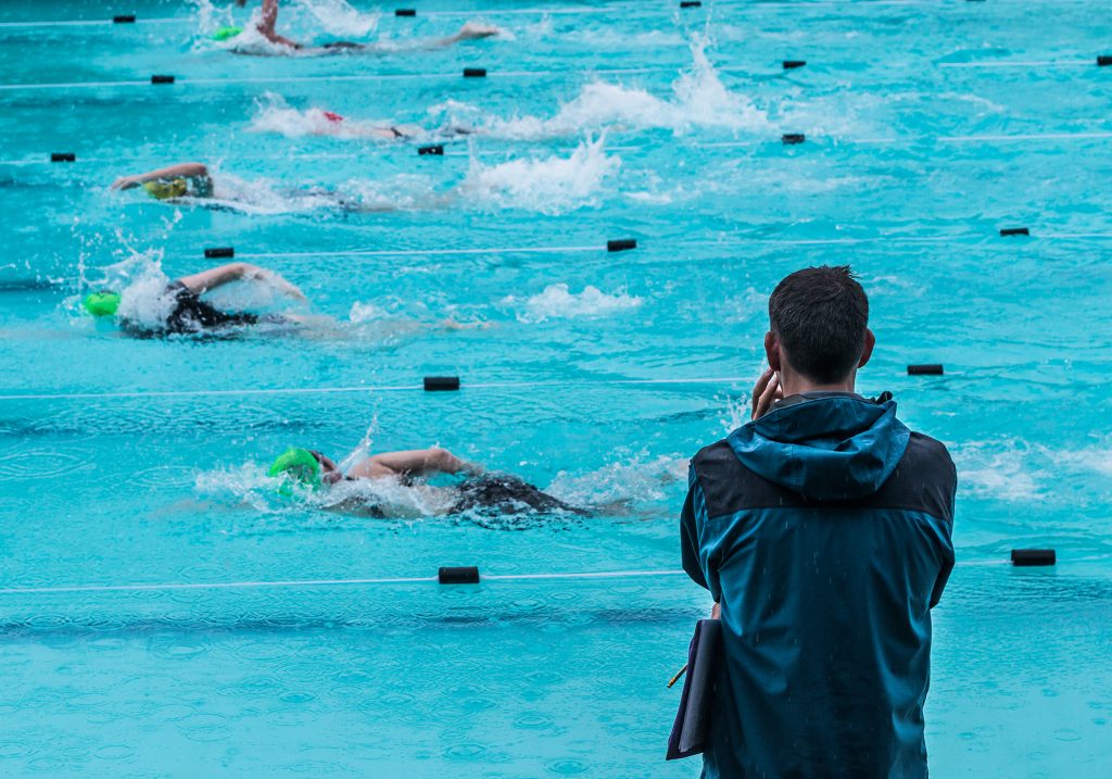 Male coach watching swimmers practice in pool lanes.